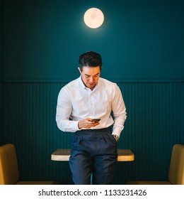 Portrait of young and attractive Chinese Asian business man using his smartphone and smiling. He is professionally dressed in a fitting shirt and pants in the office. 
