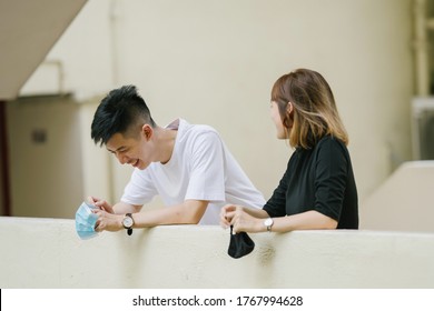 Portrait of a young and attractive Chinese Asian couple as they lean against a railing on a balcony and talk to one another. They are both relaxed and happy and are holding onto face masks.  - Powered by Shutterstock