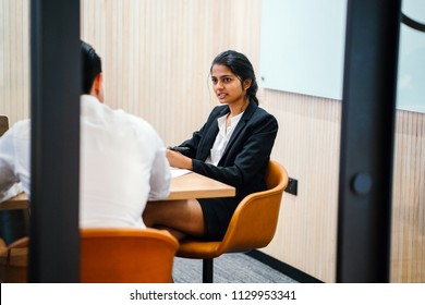 Portrait Of A Young, Attractive And Confident Indian Asian Woman Having A Meeting With A Chinese Man In A Meeting Room. She Is Either Having An Interview Or Is Giving A Performance Review. 