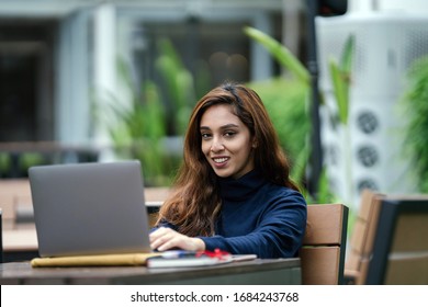 Portrait of a young, attractive, confident, smart and studious Indian Asian smiling as she types and works on her laptop computer. She is sitting at a desk in her university, office or coworking space