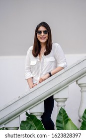 Portrait Of A Young, Attractive And Confident Thai Asian Woman In A Casual Outfit And Sunglasses Smiling As She Relaxes And Leans On The Railing Of The Stairs Of An Old Colonial Building In The Day.