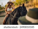 Portrait of young attractive cowgirl with cowboy hat horseback riding at homestead. Happy handsome rider in western styled outfit is riding a horse at horse ranch. Young horsewoman with stetson hat.