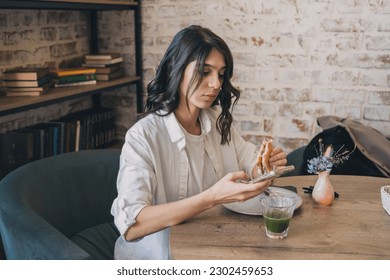 Portrait of a young attractive dark-haired woman eating a sandwich in a cafe and using a smartphone.Remote work,business, freelance,blogging,social media concept. - Shutterstock ID 2302459653