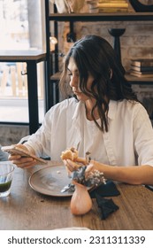 Portrait of a young attractive dark-haired woman eating a sandwich in a cafe and using a smartphone.Remote work,business, freelance,blogging,social media concept. - Shutterstock ID 2311311339