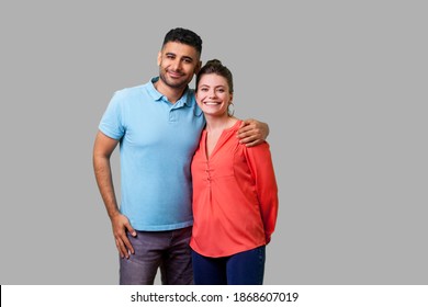 Portrait Of Young Attractive Family Couple In Casual Wear Standing Together, Embracing And Looking At Camera With Sincere Smile, Strong Relations. Isolated On Gray Background, Indoor Studio Shot