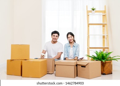 Portrait Of Young Attractive Happy Asian Couple Man And Woman Sitting At New Home Smiling To Camera With Carton Package Box Storage To Move In Empty House. Young Married Asian Relocating Home Concept.