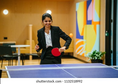 Portrait of a young and attractive Indian Asian woman in a suit playing table tennis with her colleague in the office during a break. She is having lots of fun and is smiling broadly. 