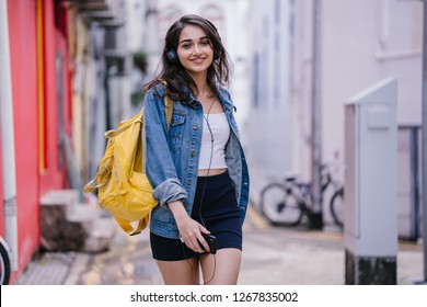 Portrait of a young and attractive Indian Asian woman in a denim jacket and yellow backpack enjoying music on her headphones. She is smiling as dancing as she walks down an alley on her way somewhere.