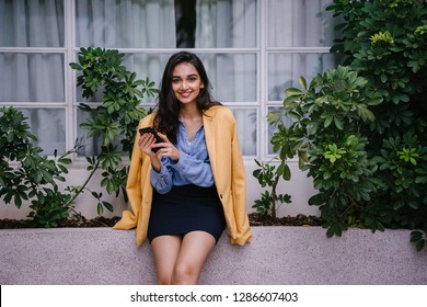 Portrait Of A Young And Attractive Indian Asian Fashionista Woman Using Her Smartphone During The Day. She Is Wearing A Vintage Yellow Jacket Draped Over A Casual Outfit And Is Smiling Happily. 