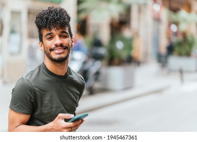 Portrait Of A Young And Attractive Latin American Man Looking At Camera And Smiling. He Is Holding A Blue Cell Phone. He Is At The Street In Barcelona.