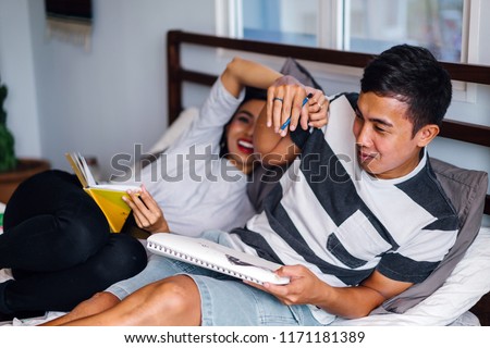 Portrait of a young and attractive Malay Asian couple sitting around lazily in bed on the weekend. The woman is tickling the man and he is laughing out loud in a shared tender moment together.