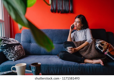 Portrait Of A Young And Attractive Malay Muslim Woman Talking On Her Smartphone. She Is Sitting On A Beautiful Blue Couch In A Colorful, Trendy And Modern Living Room. 