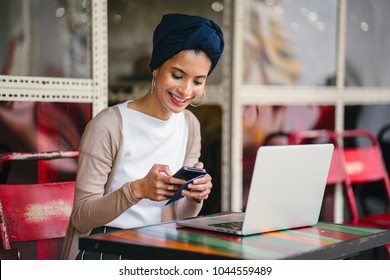 Portrait Of A Young, Attractive Muslim Woman (Asian, Arab, Malay) Entrepreneur Or Businesswoman Working On Her Smartphone. She Is Dressed Smartly And Wears A Turban (hijab, Headscarf) And Is Sitting. 