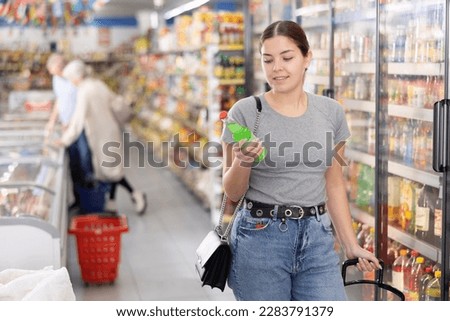 Portrait of young attractive positive girl walking down aisle of grocery store, choosing refreshing non-alcoholic beverages