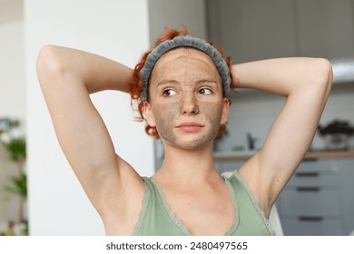 portrait young attractive positive woman, red-haired, curly, with bandage on hair, morning in cosmetic mask, in kitchen, stretching contentedly, raising hands up - Powered by Shutterstock