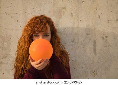Portrait of young, attractive, red-haired, freckled, red-sweatered woman filling an orange globe on a gray wall. Concept colors, balloons, beauty, fashion, red.