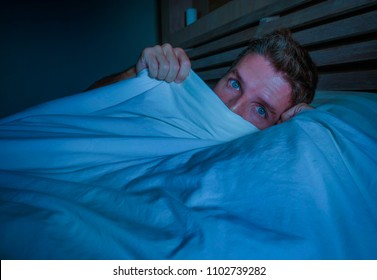 Portrait Of Young Attractive Scared Man In Fear And Panic Suffering Horror Nightmare Covering Face With Blanket Sleepless At Night Lying On Bed In Dark Bedroom In Sleeping Disorder Concept