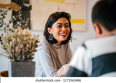 Portrait Of A Young And Attractive Singaporean Malay Woman Enjoying A Hot Coffee Beverage While She Talks To A Man In A Living Room During The Day. She Is Smiling As She Talks. 