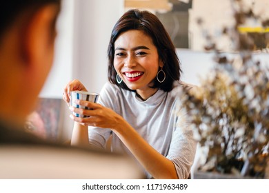 Portrait Of A Young And Attractive Singaporean Malay Woman Enjoying A Hot Coffee Beverage While She Talks To A Man In A Living Room During The Day. She Is Smiling As She Talks. 