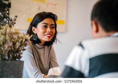 Portrait Of A Young And Attractive Singaporean Malay Woman Enjoying A Hot Coffee Beverage While She Talks To A Man In A Living Room During The Day. She Is Smiling As She Talks. 