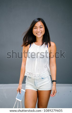 Portrait of a young, attractive and tanned Chinese Asian woman against a grey background as she uses her smartphone. She is smiling in a candid way as she taps on her phone. 