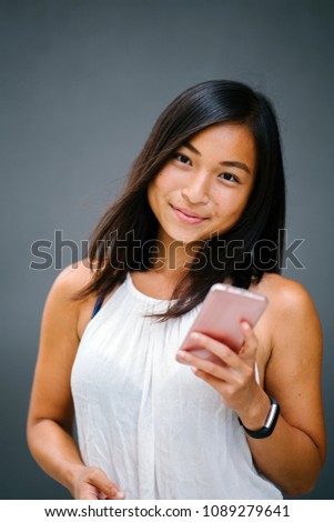 Portrait of a young, attractive and tanned Chinese Asian woman against a grey background as she uses her smartphone. She is smiling in a candid way as she taps on her phone. 