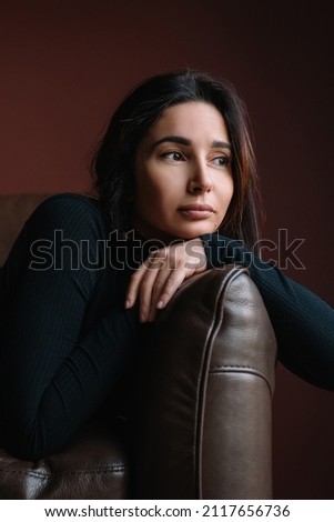 Portrait of young attractive thoughtful woman with long dark hair in black roll-neck sit on brown sofa putting head on crossed arms. Depression, fatigue, stress, problems, emotions concept. Vertical. 