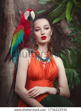 Portrait of young attractive woman in african style with ara parrot on her hand on a tropical background. Beautiful brunette girl in drungles with a macaw parrot. Artistic processing.