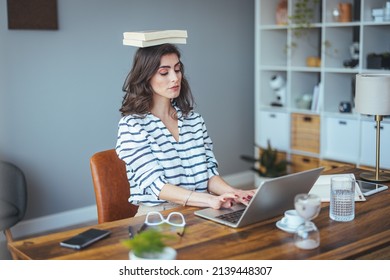 Portrait of a young attractive woman at the desk with books on her head, sitting straight, working on laptop. Education concept photo, lifestyle. 