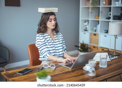 Portrait of a young attractive woman at the desk with books on her head, sitting straight, working on laptop. Education concept photo, lifestyle. 