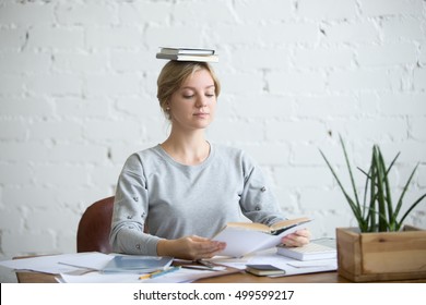 Portrait of a young attractive woman at the desk with books on her head, sitting straight, reading a book. Education concept photo, lifestyle