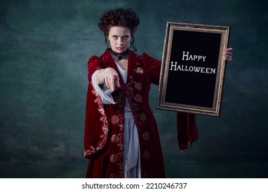 Portrait Of Young Attractive Woman In Image Of Vampire Holding Picture Frame Isolated Over Dark Green Background. Century Of Monsters. Concept Of Halloween, Mystery, Gothic Art, Fantasy, Surreal