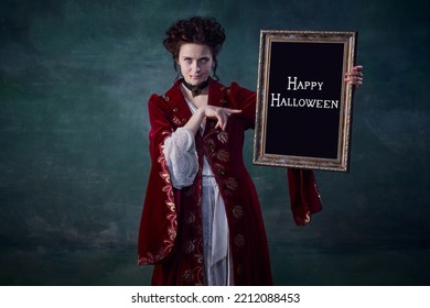 Portrait Of Young Attractive Woman In Image Of Vampire Holding Picture Frame Isolated Over Dark Green Background. Horror Time. Concept Of Halloween, Mystery, Gothic Art, Fantasy, Surreal Characters