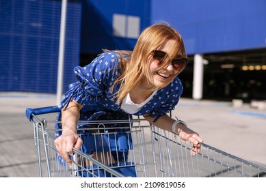 Portrait Of A Young Attractive Woman In An Outdoor Supermarket Parking In A Trolley. Shopping, Fun And Shopping Concept