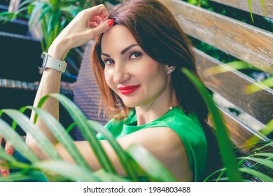 Portrait of young attractive woman with red hair and bright red lips in green short sleeve blouse. Woman is sitting on wooden bench among green plants and smiling. Selective soft focus, toned image
