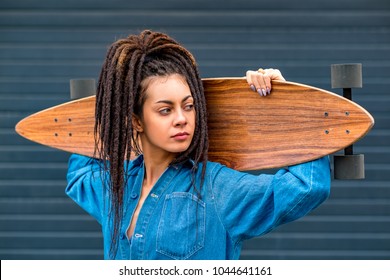 Portrait of young attractive woman or teenage girl in blue denim romper shirt with dreadlocks holding wooden longboard skateboard on her shoulders. Urban scene, city life. Hipster cute lady. - Powered by Shutterstock