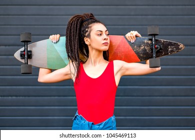 Portrait of a young attractive woman or teenage girl with dreadlocks holding wooden longboard skateboard in front of her. Urban scene, city life. Hipster cute lady.