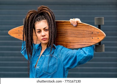 Portrait of young attractive woman or teenage girl in blue denim overall shirt with dreadlocks holding wooden longboard skateboard on her shoulders. Urban scene, city life. Hipster cute lady. - Powered by Shutterstock