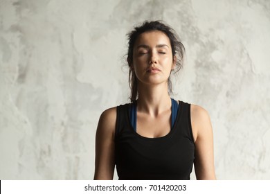 Portrait of young attractive yogi woman with her eyes closed in meditating pose, relaxation exercise, working out, wearing sportswear, black top, indoor close up image, wall background