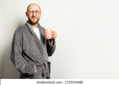 Portrait Of A Young Bald Man With A Beard In A Dressing Gown And Glasses Holding A Cup Of Hot Coffee Or Tea On Light Background. Emotional Face. Concept Of Morning Coffee, Morning, Evening Coffee