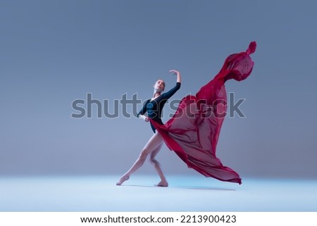 Portrait of young ballerina dancing in black bodysuit with deep red fabric isolated on blue grey studio background. Grace, tenderness. Concept of classic ballet, inspiration, beauty, dance, creativity