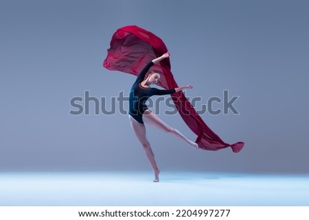 Portrait of young ballerina dancing with deep red fabric isolated over blue grey studio background. Tender wave. Concept of classic ballet, inspiration, beauty, dance, creativity