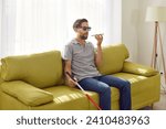 Portrait of a young bearded blind man in black eyeglasses holding cane and using digital assistant on mobile phone sitting on sofa at home. Visually impaired person talking on mobile phone.