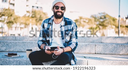 Portrait of young bearded male blogger with palmtop technology looking at camera listening music podcast during travel vacations, Middle Eastern hipster guy in earphones holding digital tablet