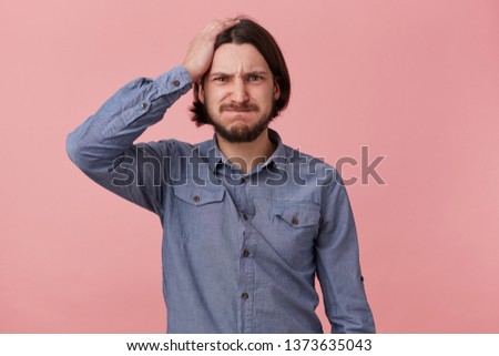 Portrait of young bearded man in denim shirt, hold his head, blowing cheeks and bites lips, forgot something important, made a mistake. Isolated over oink background.