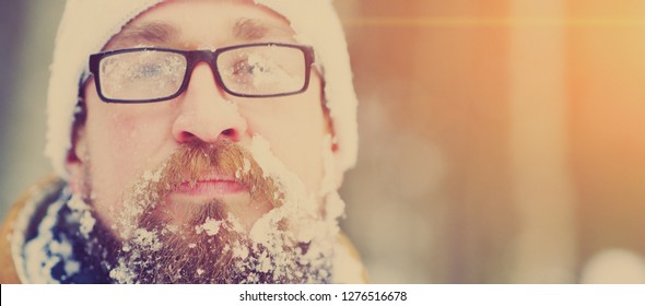 Portrait of a young bearded man frozen in a Blizzard in the woods. Face, beard , mustache and glasses covered with frost. Face covered with snow.