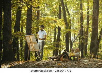 Portrait of young bearded man in hat painting a picture using brushes and modern easel on autumn nature in a autumnal park next to his wife and son. Painter artist forest. Art concept. - Powered by Shutterstock