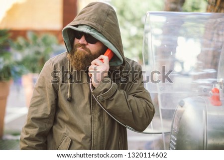 Portrait of young bearded man using public phone wearing hoodie (jacket) and black sunglasses - informant, spy, secret agent and fake news concept