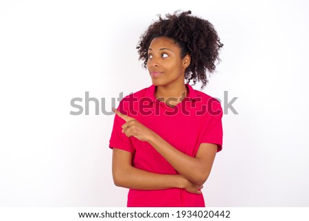 Portrait of young beautiful African American woman wearing pink t-shirt against white wall posing on camera with tricky look, presenting product with index finger. Advertisement concept.