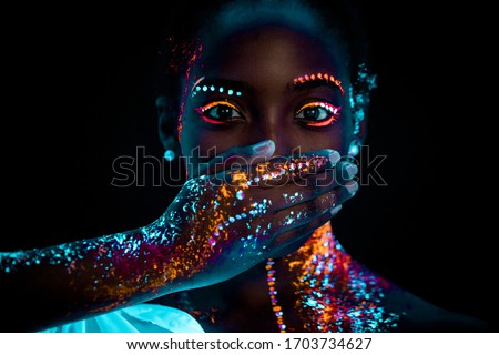 portrait of young beautiful african woman with fluorescent prints on face and closed mouth. fluorescent prints glowing on ultra violet rays, keep silence gesture